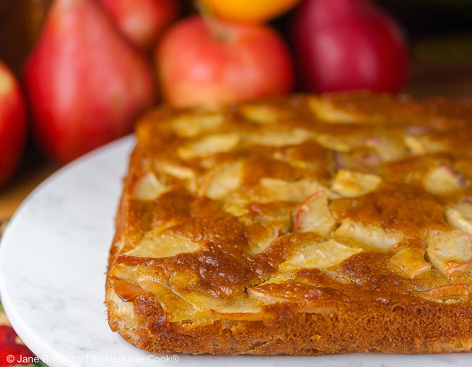 A gorgeous browned square cake with tons of fruit breaking the surface on a round white marble surface with red and orange fall fruits behind, a slice with a sprig of mint and red grapes flanking it © 2026 Jane Bonacci, The Heritage Cook.