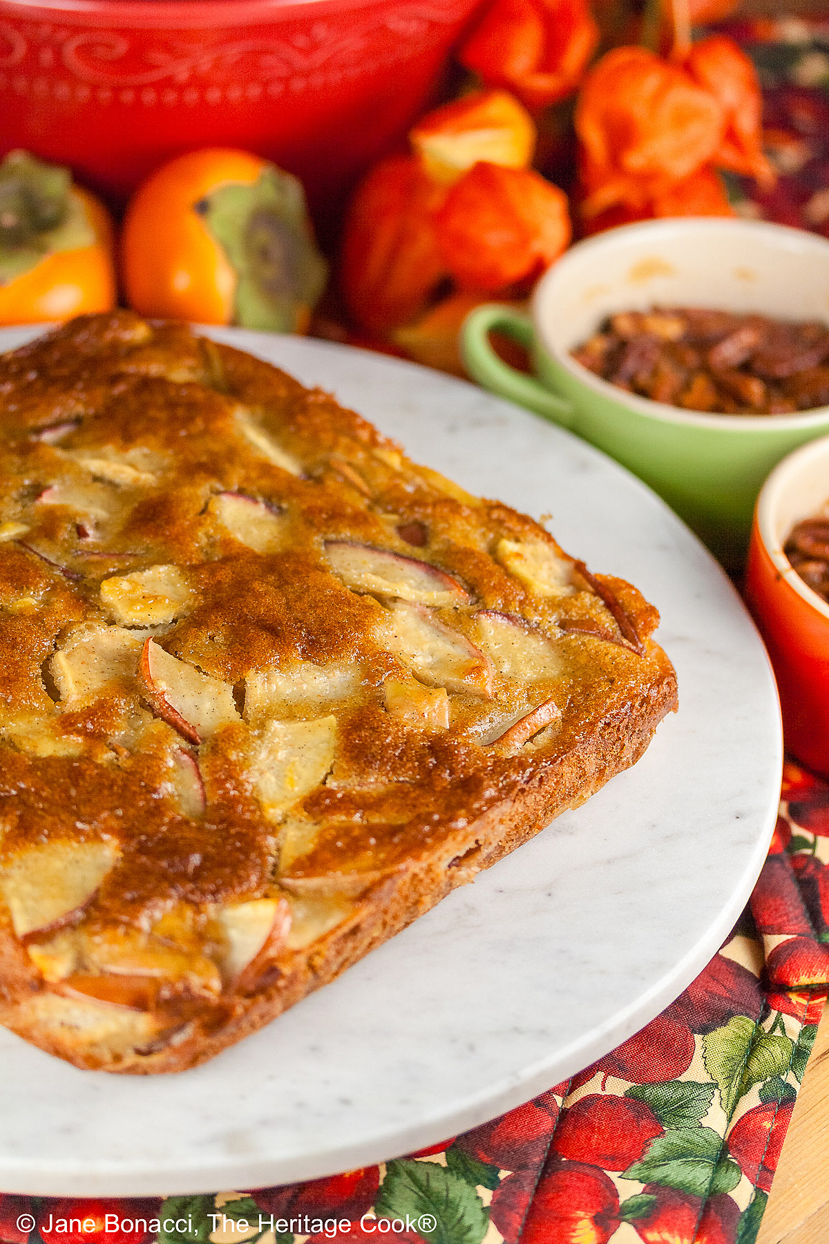 A gorgeous browned square cake with tons of fruit breaking the surface on a round white marble surface with red and orange fall fruits behind, a slice with a sprig of mint and red grapes flanking it © 2026 Jane Bonacci, The Heritage Cook.