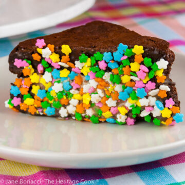 Triangular Brownie Ice Cream Sandwiches with the edges dipped in a variety of items, like rainbow sprinkles, coconut, and chocolate chunks stacked on a white marble turntable in front of a bright orange pitcher, with a white plate in front with a single ice cream sandwich dipped in rainbow sprinkles on it, sitting on a brightly colored pastel checked cloth © 2026 Jane Bonacci, The Heritage Cook.