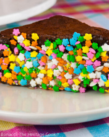 Triangular Brownie Ice Cream Sandwiches with the edges dipped in a variety of items, like rainbow sprinkles, coconut, and chocolate chunks stacked on a white marble turntable in front of a bright orange pitcher, with a white plate in front with a single ice cream sandwich dipped in rainbow sprinkles on it, sitting on a brightly colored pastel checked cloth © 2026 Jane Bonacci, The Heritage Cook.