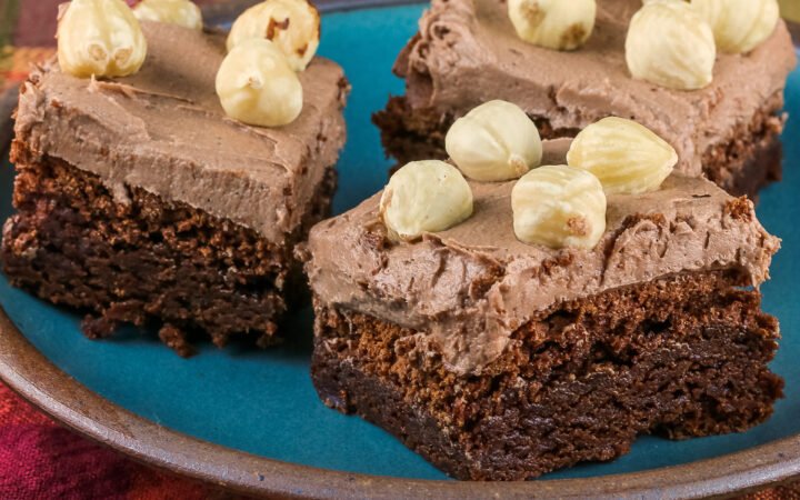 3 brownies on a blue plate, topped with Nutella frosting and studded with whole hazelnuts; Brownies with Nutella Frosting © 2022 Jane Bonacci, The Heritage Cook.