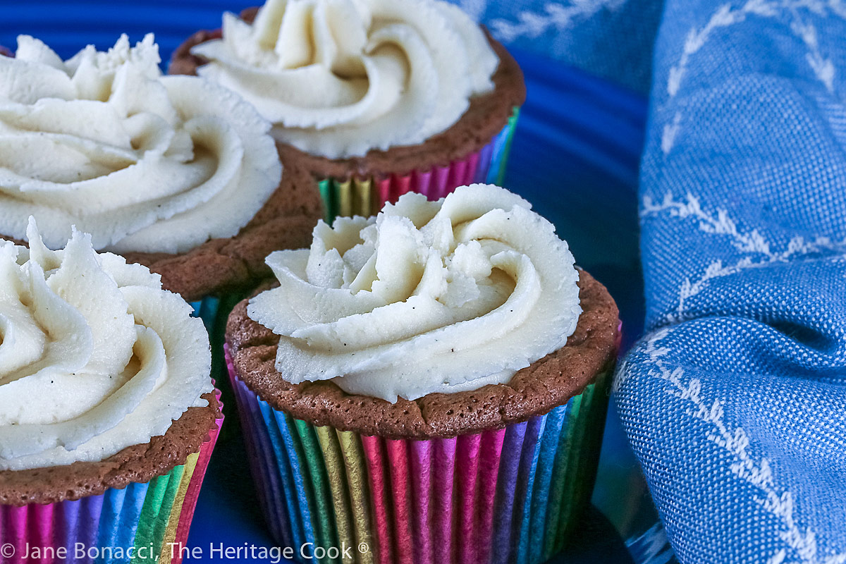 Chocolate cupcakes in rainbow colored cupcake liner papers on a blue plate with a blue and white cloth; Chocolate Velvet Cupcakes topped with Cream Cheese Frosting © 2022 Jane Bonacci, The Heritage Cook.