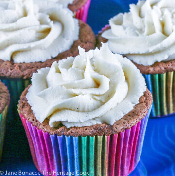 Chocolate cupcakes in rainbow colored cupcake liner papers on a blue plate with a blue and white cloth; Chocolate Velvet Cupcakes topped with Cream Cheese Frosting © 2022 Jane Bonacci, The Heritage Cook.