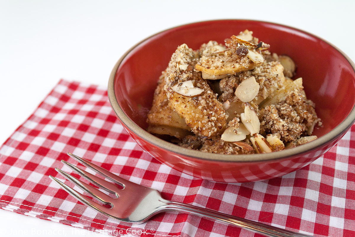 Sweetened apple filling and crispy topping with almonds served in a red bowl; Gluten-Free Apple Crisp © 2022 Jane Bonacci, The Heritage Cook.