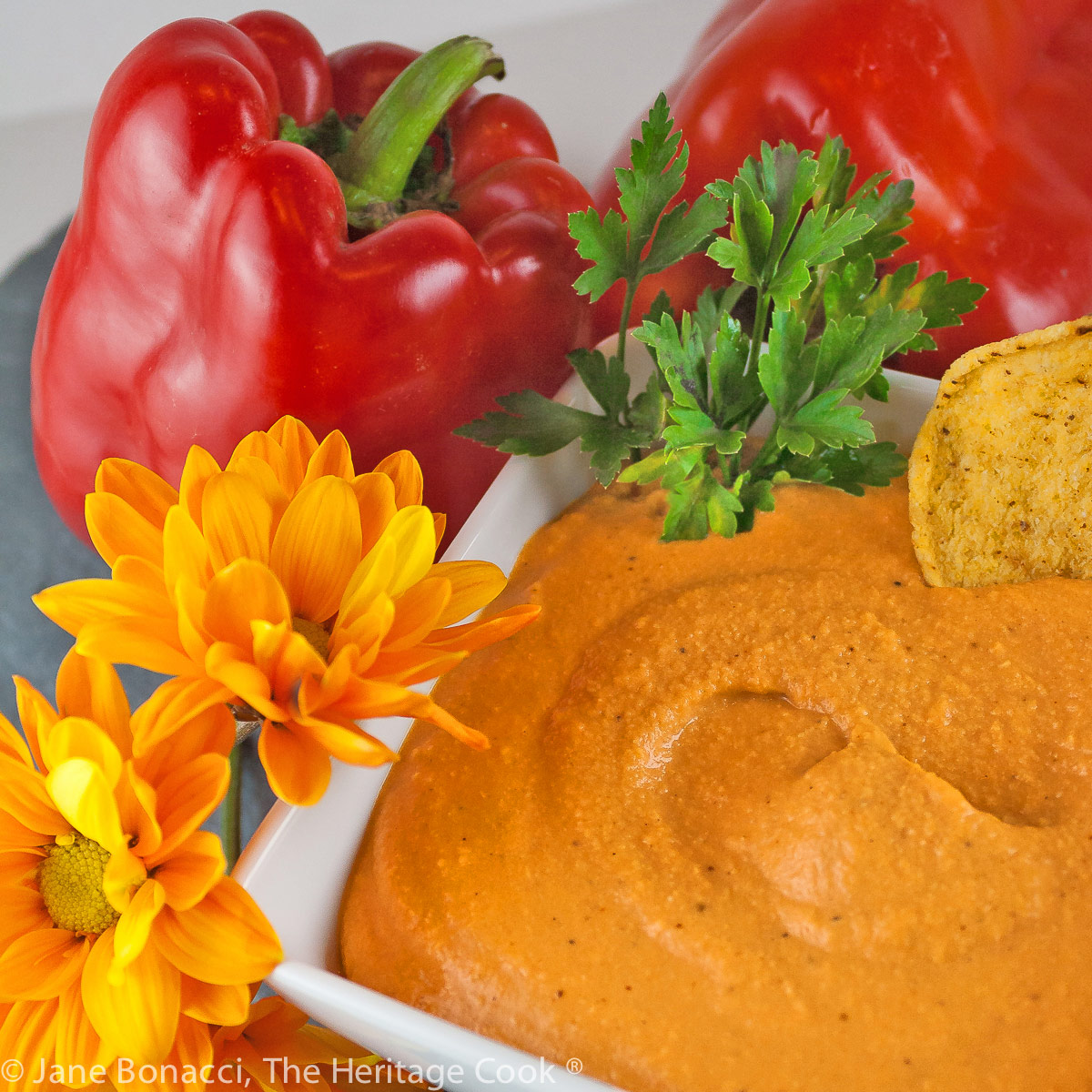 Orange roasted red pepper hummus in a square white bowl surrounded by red bell peppers, tortilla chips, parsley, and yellow fall flowers; © 2022 Jane Bonacci, The Heritage Cook.