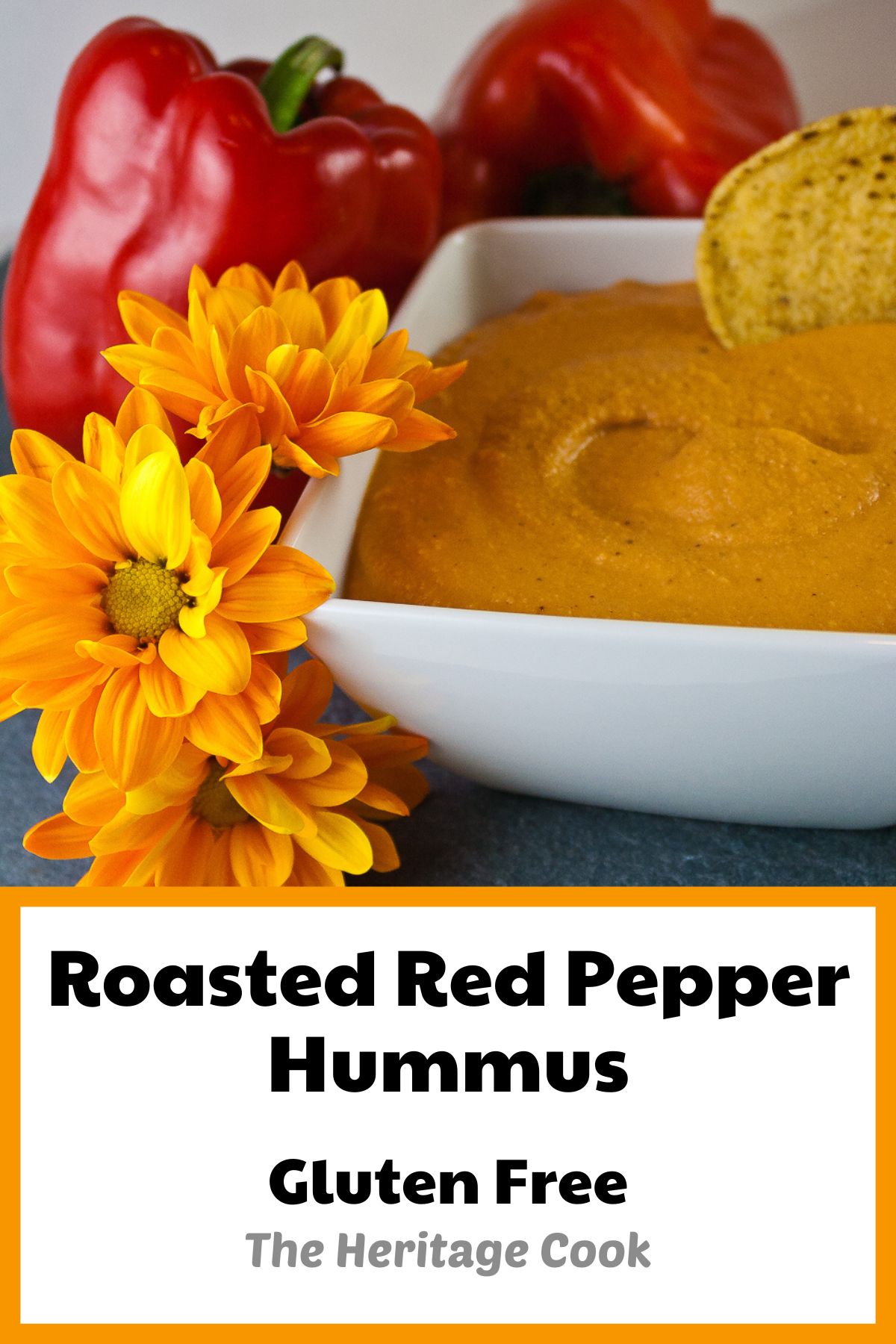 Orange roasted red pepper hummus in a square white bowl surrounded by red bell peppers, tortilla chips, parsley, and yellow fall flowers; © 2022 Jane Bonacci, The Heritage Cook.