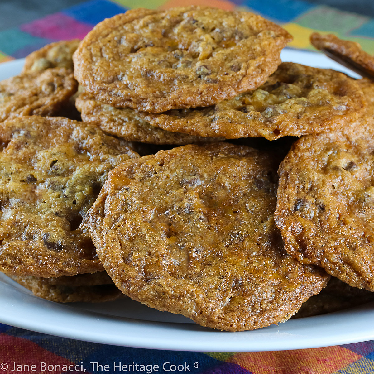 Pile of baked cookies on a white plate with a brightly colored plaid cloth underneath. Easy Toffee Chocolate Chip Cookies (Gluten Free) © 2022 Jane Bonacci, The Heritage Cook. 