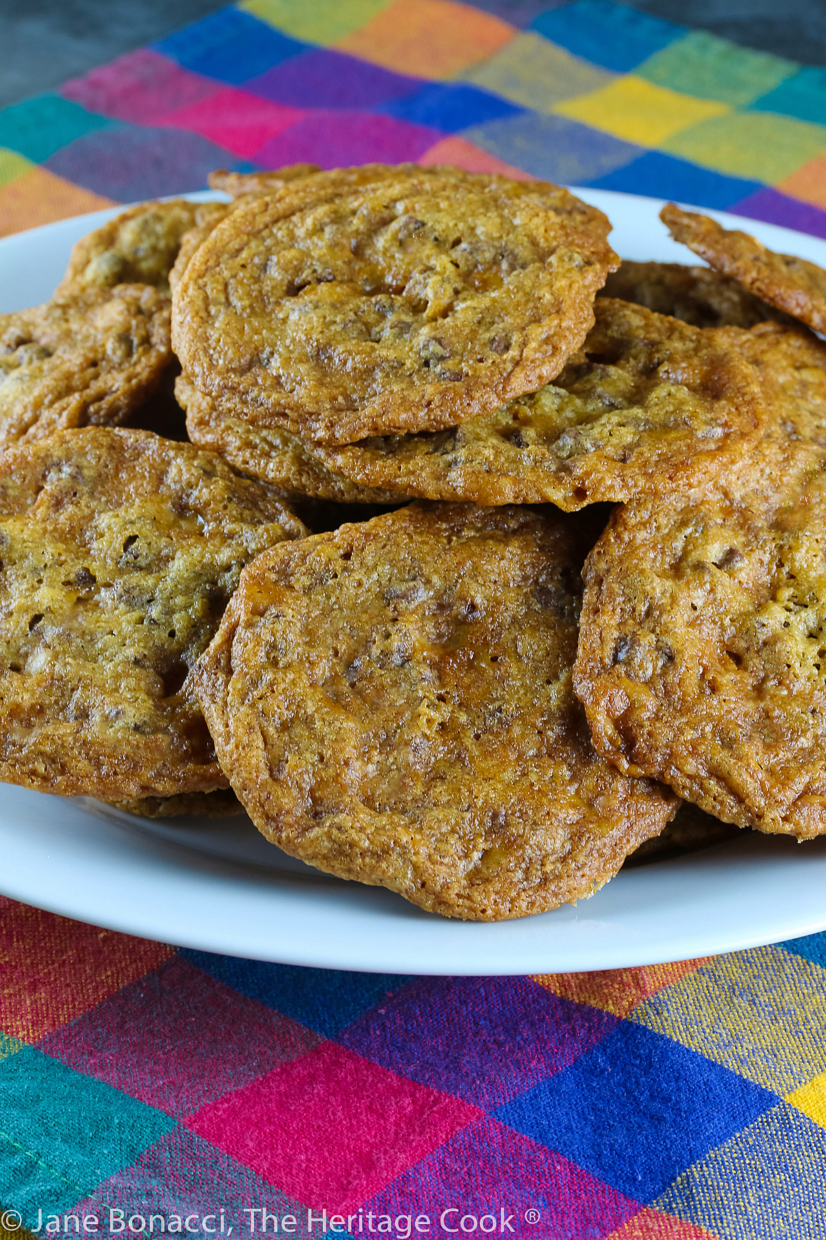 Pile of baked cookies on a white plate with a brightly colored plaid cloth underneath. Easy Toffee Chocolate Chip Cookies (Gluten Free) © 2022 Jane Bonacci, The Heritage Cook. 