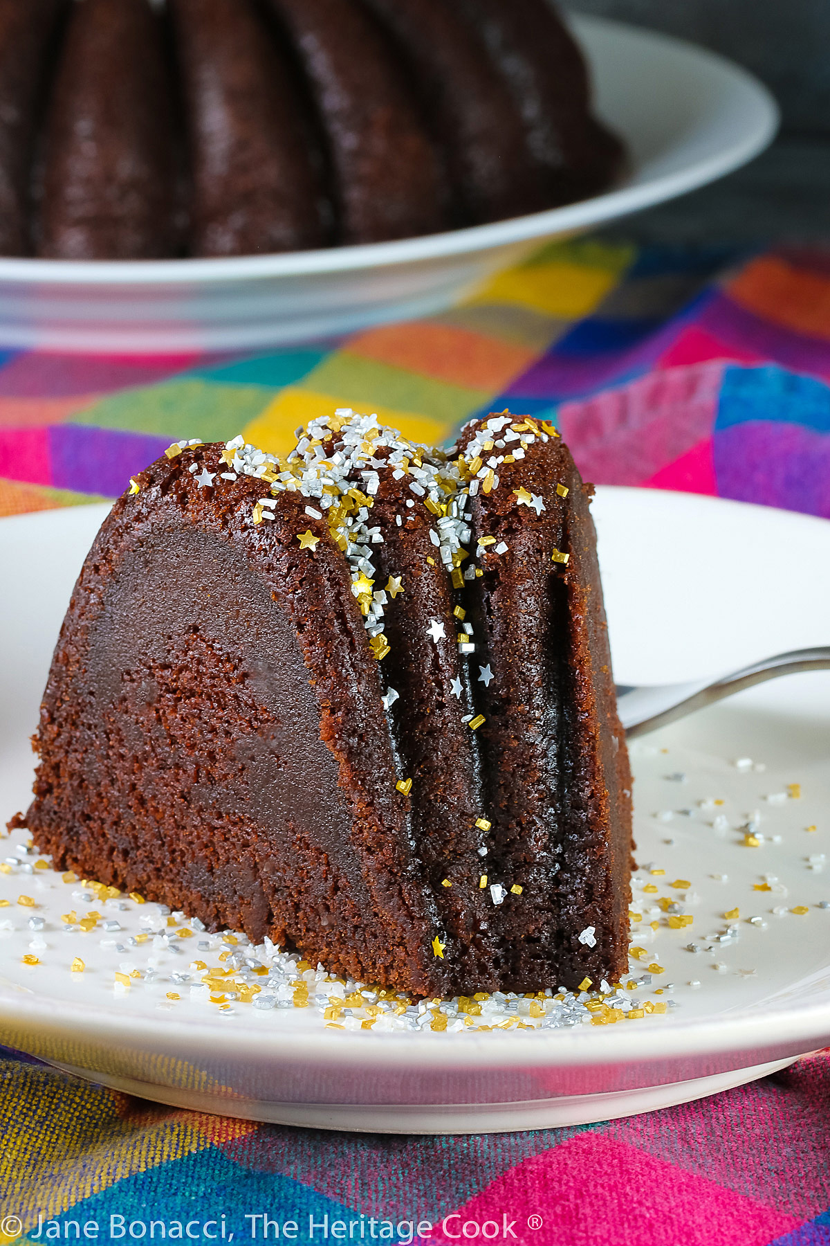 Slice of chocolate bundt cake on white plate in front of the remaining whole cake, topped with silver and gold sprinkles like a snow shower; Chocolate Bourbon Pound Cake © 2023 Jane Bonacci, The Heritage Cook. 