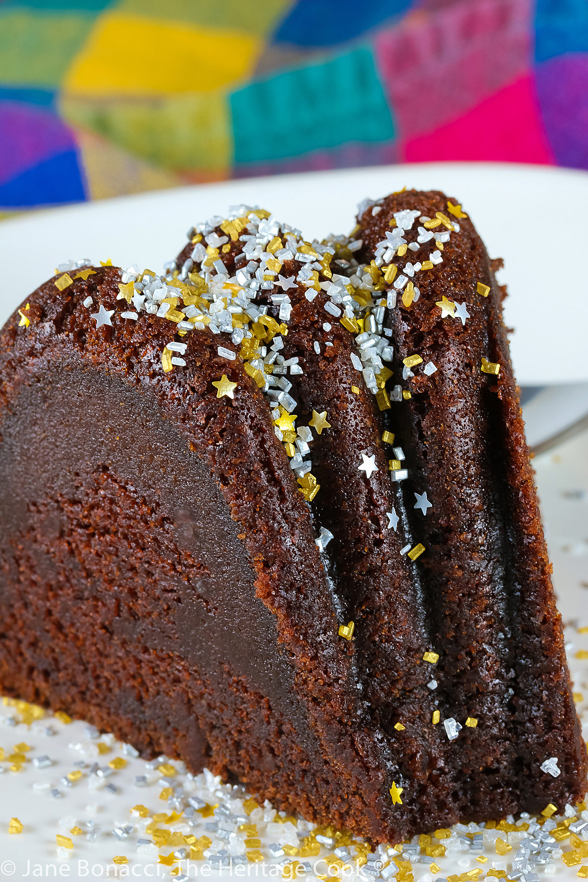 Slice of chocolate bundt cake on white plate in front of the remaining whole cake, topped with silver and gold sprinkles like a snow shower; Chocolate Bourbon Pound Cake © 2023 Jane Bonacci, The Heritage Cook. 