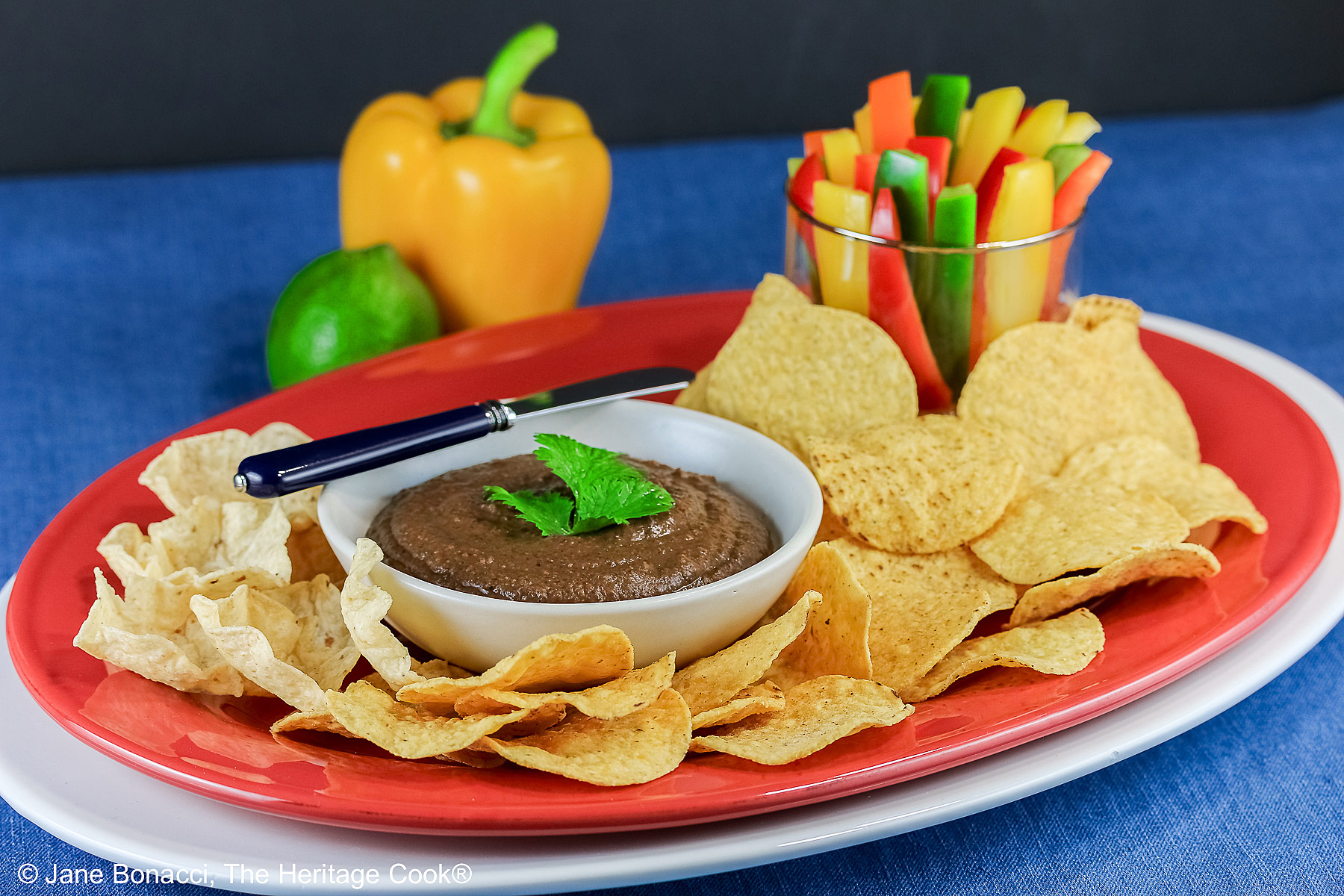 Bowl of Chipotle Black Bean Dip with a cilantro leaf on top, surrounded by yellow tortilla chips with a glass filled with bell pepper strips on one end and a whole lime and yellow bell pepper behind; sitting on a blue cloth with a black backdrop © 2026 Jane Bonacci, The Heritage Cook.
