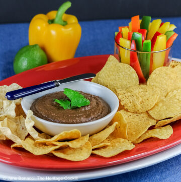Bowl of Chipotle Black Bean Dip with a cilantro leaf on top, surrounded by yellow tortilla chips with a glass filled with bell pepper strips on one end and a whole lime and yellow bell pepper behind; sitting on a blue cloth with a black backdrop © 2026 Jane Bonacci, The Heritage Cook.