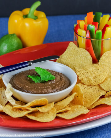 Bowl of Chipotle Black Bean Dip with a cilantro leaf on top, surrounded by yellow tortilla chips with a glass filled with bell pepper strips on one end and a whole lime and yellow bell pepper behind; sitting on a blue cloth with a black backdrop © 2026 Jane Bonacci, The Heritage Cook.