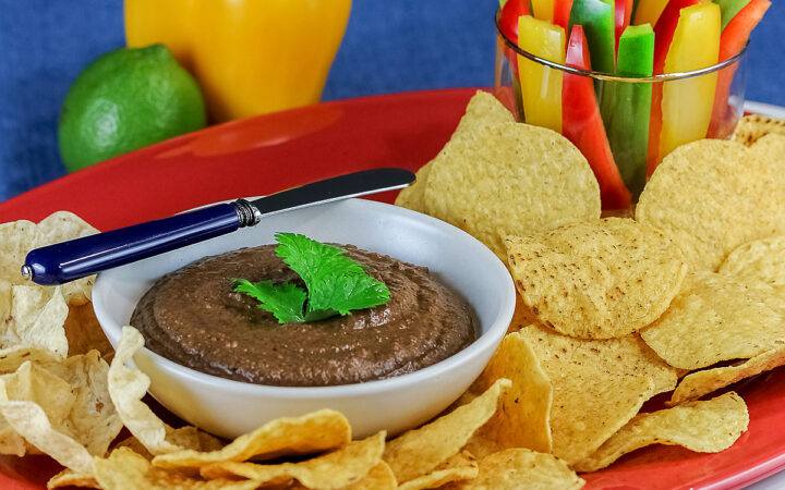 Bowl of Chipotle Black Bean Dip with a cilantro leaf on top, surrounded by yellow tortilla chips with a glass filled with bell pepper strips on one end and a whole lime and yellow bell pepper behind; sitting on a blue cloth with a black backdrop © 2026 Jane Bonacci, The Heritage Cook.