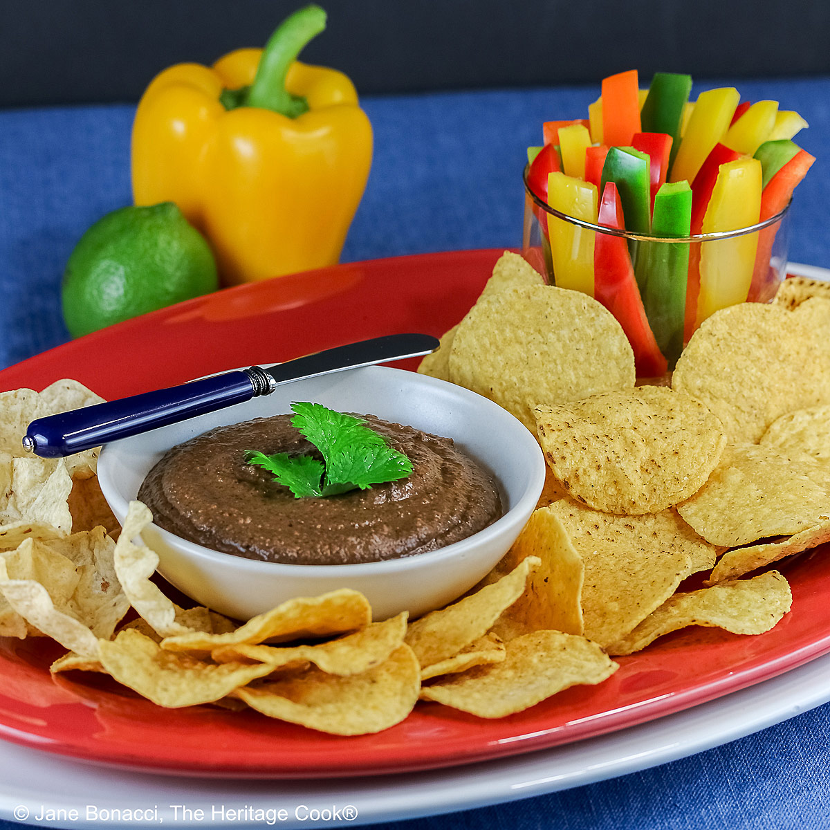 Bowl of Chipotle Black Bean Dip with a cilantro leaf on top, surrounded by yellow tortilla chips with a glass filled with bell pepper strips on one end and a whole lime and yellow bell pepper behind; sitting on a blue cloth with a black backdrop © 2026 Jane Bonacci, The Heritage Cook.