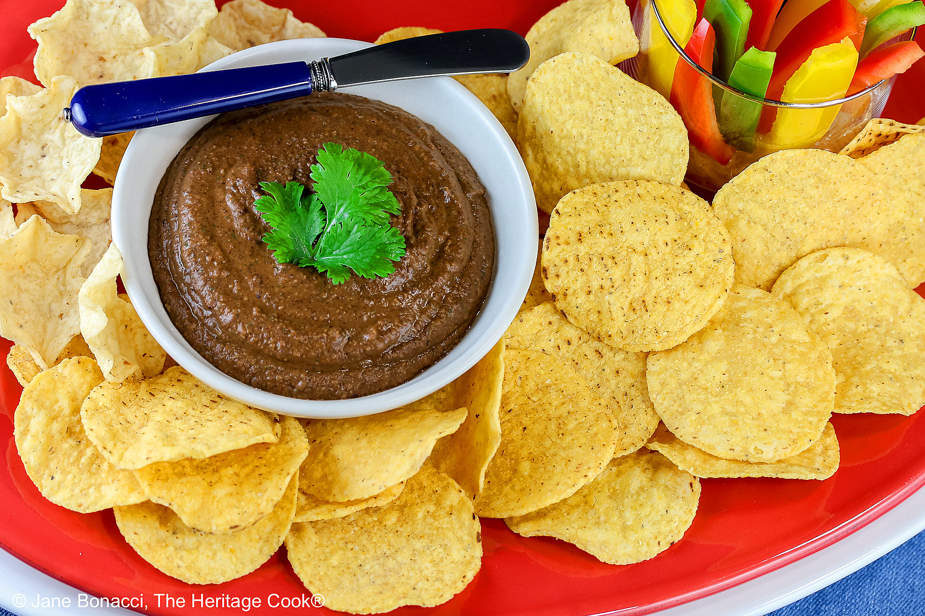 Bowl of Chipotle Black Bean Dip with a cilantro leaf on top, surrounded by yellow tortilla chips with a glass filled with bell pepper strips on one end and a whole lime and yellow bell pepper behind; sitting on a blue cloth with a black backdrop © 2026 Jane Bonacci, The Heritage Cook.
