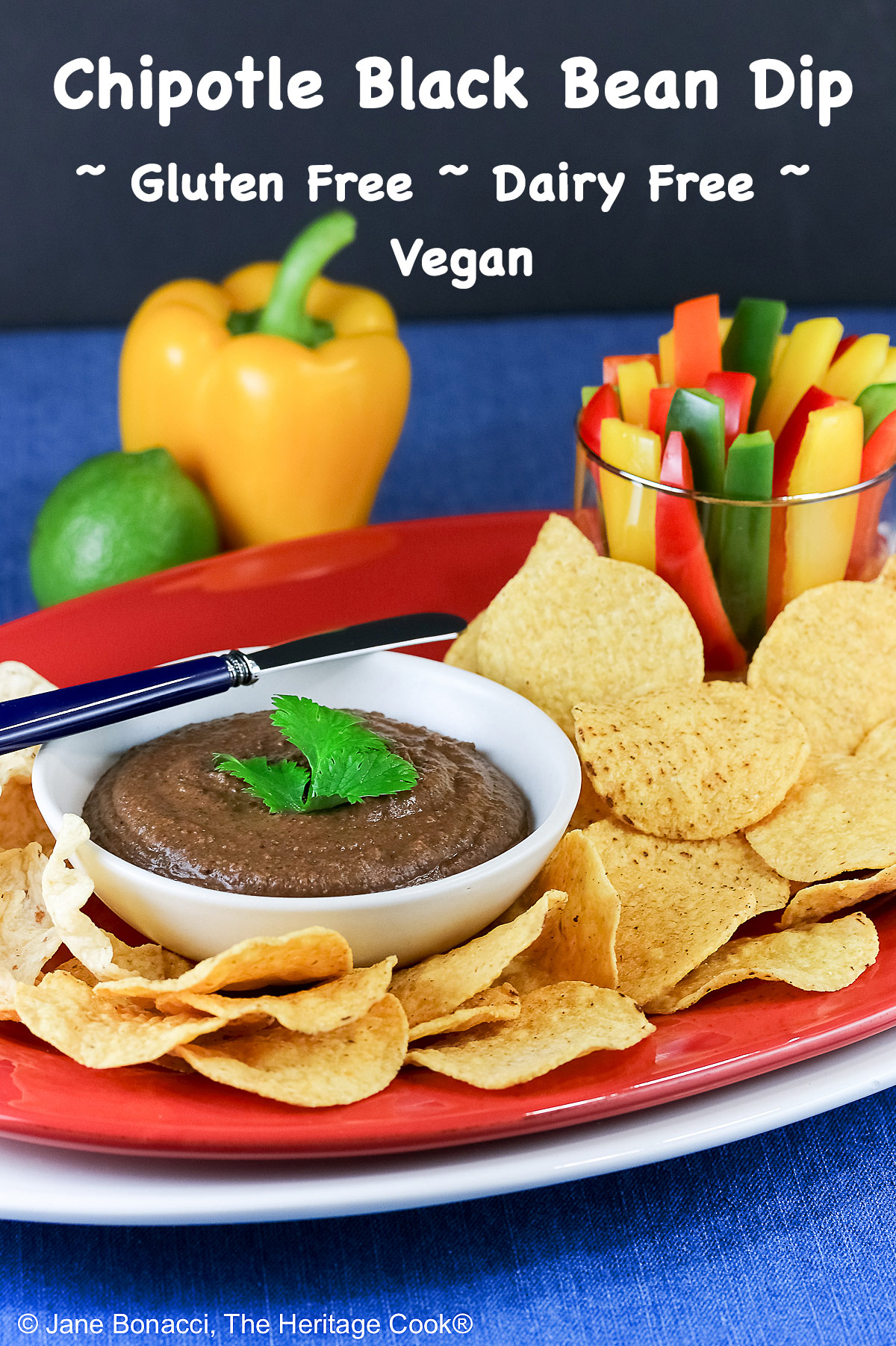 Bowl of Chipotle Black Bean Dip with a cilantro leaf on top, surrounded by yellow tortilla chips with a glass filled with bell pepper strips on one end and a whole lime and yellow bell pepper behind; sitting on a blue cloth with a black backdrop © 2026 Jane Bonacci, The Heritage Cook.