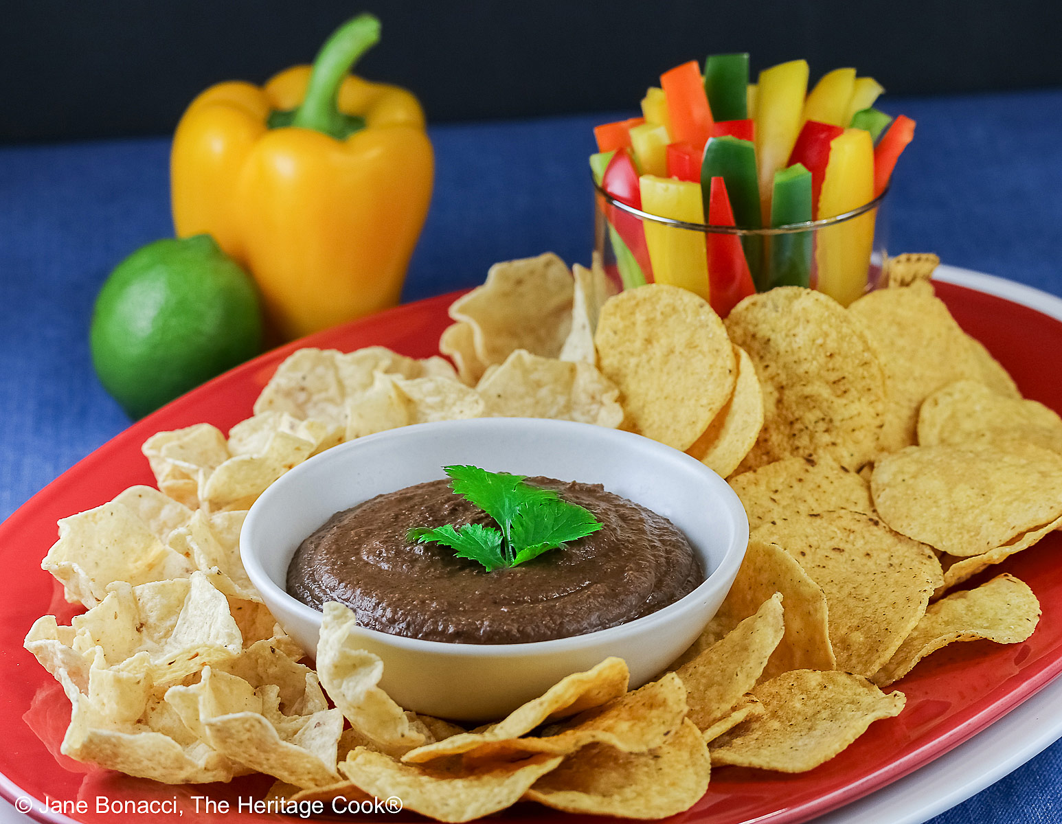 Bowl of Chipotle Black Bean Dip with a cilantro leaf on top, surrounded by yellow tortilla chips with a glass filled with bell pepper strips on one end and a whole lime and yellow bell pepper behind; sitting on a blue cloth with a black backdrop © 2026 Jane Bonacci, The Heritage Cook.