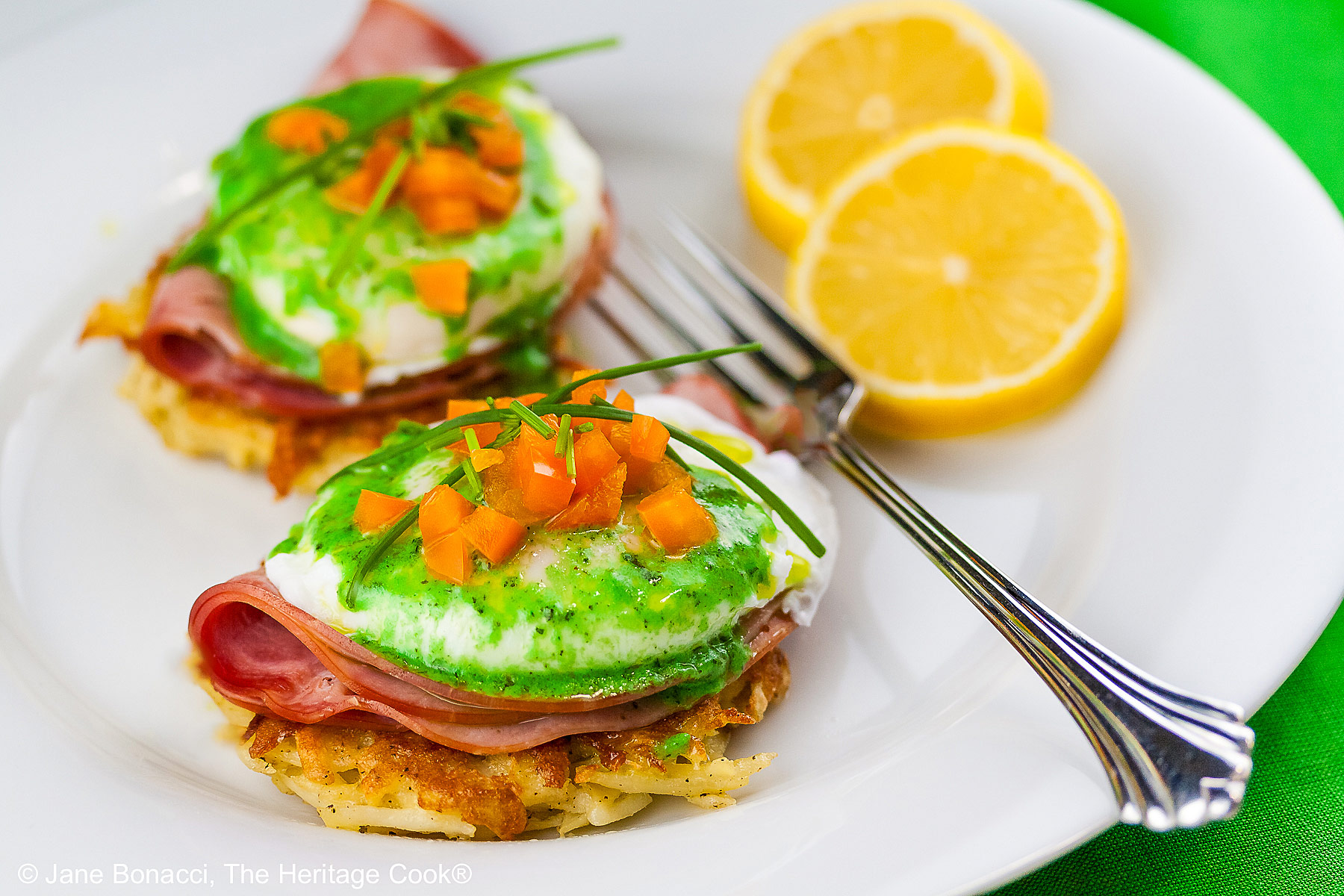 Two beautifully poached eggs laid on top of a slice of ham on a fried hash brown patty and topped with bright green hollandaise sauce, orange bell pepper cubes, and chives to mimic the colors of the Irish flag, on a white plate with two slices of lemon © 2026 Jane Bonacci, The Heritage Cook.