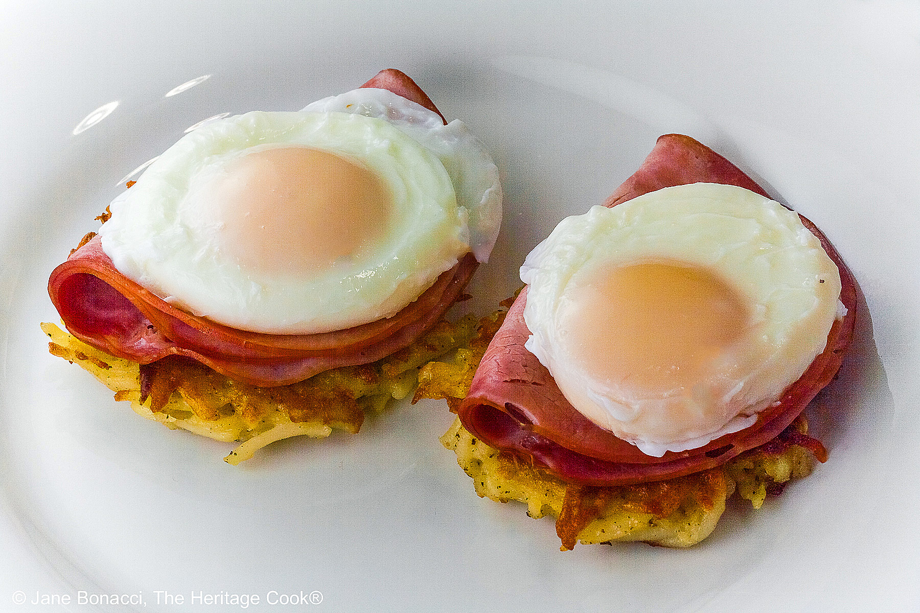 2 perfectly poached eggs sitting on a slice of ham and a fried potato patty.