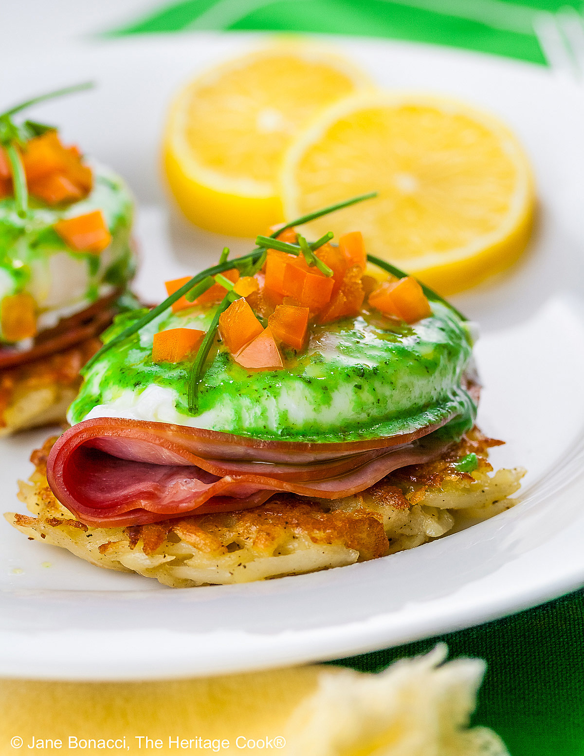 Two beautifully poached eggs laid on top of a slice of ham on a fried hash brown patty and topped with bright green hollandaise sauce, orange bell pepper cubes, and chives to mimic the colors of the Irish flag, on a white plate with two slices of lemon © 2026 Jane Bonacci, The Heritage Cook.
