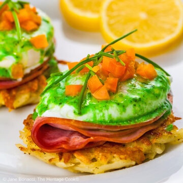 Two beautifully poached eggs laid on top of a slice of ham on a fried hash brown patty and topped with bright green hollandaise sauce, orange bell pepper cubes, and chives to mimic the colors of the Irish flag, on a white plate with two slices of lemon © 2026 Jane Bonacci, The Heritage Cook.