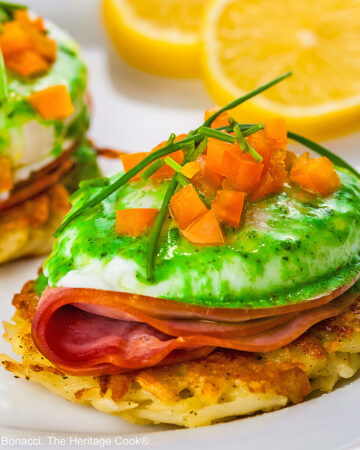 Two beautifully poached eggs laid on top of a slice of ham on a fried hash brown patty and topped with bright green hollandaise sauce, orange bell pepper cubes, and chives to mimic the colors of the Irish flag, on a white plate with two slices of lemon © 2026 Jane Bonacci, The Heritage Cook.