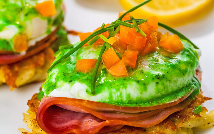 Two beautifully poached eggs laid on top of a slice of ham on a fried hash brown patty and topped with bright green hollandaise sauce, orange bell pepper cubes, and chives to mimic the colors of the Irish flag, on a white plate with two slices of lemon © 2026 Jane Bonacci, The Heritage Cook.