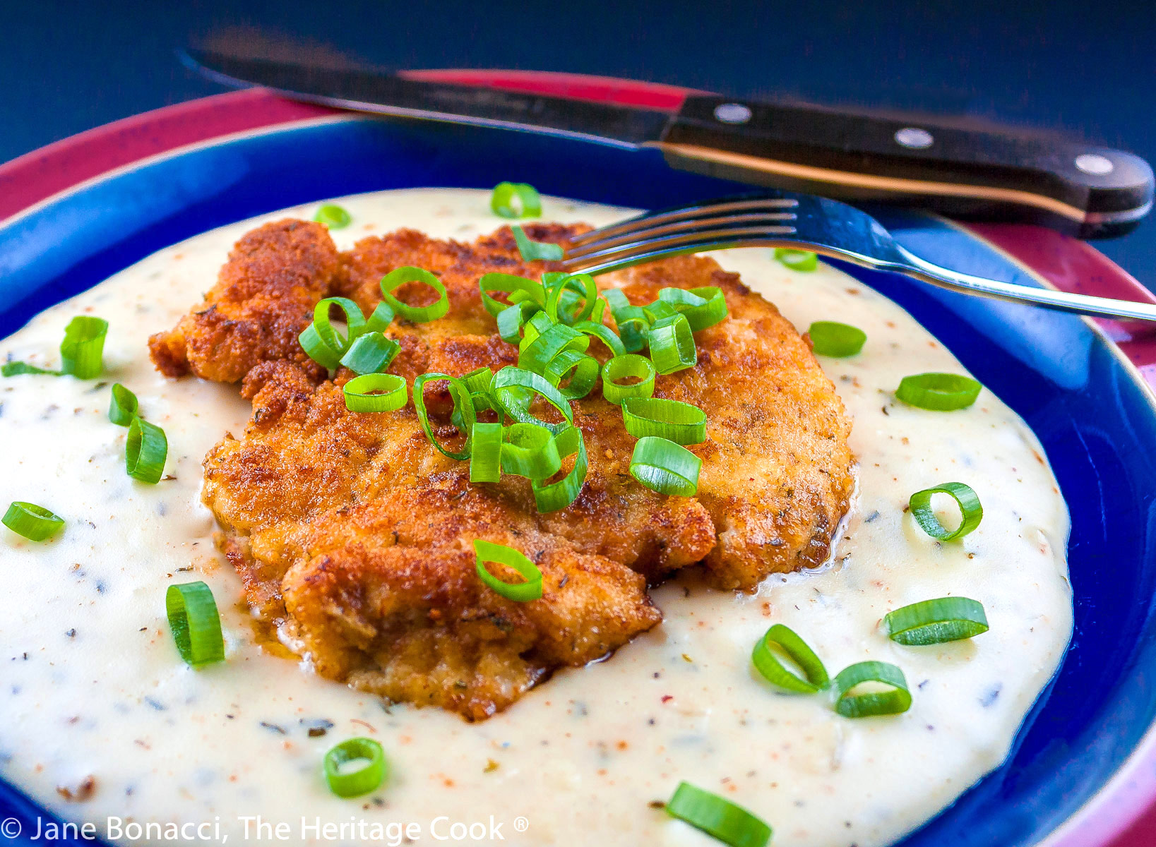 Golden fried chicken breasts resting on cream gravy and topped with green onions on blue and red plate; Fried Chicken Cutlets with Cream Gravy © 2023 Jane Bonacci, The Heritage Cook. 