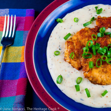Golden fried chicken breasts resting on cream gravy and topped with green onions on blue and red plate; Fried Chicken Cutlets with Cream Gravy © 2023 Jane Bonacci, The Heritage Cook.