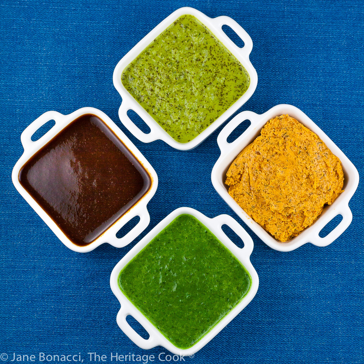 Mini white containers with four sauces for BBQ foods, two herbal sauces, a BBQ sauce, and a compound butter, all on a bright blue background; © 2023 Jane Bonacci, The Heritage Cook.