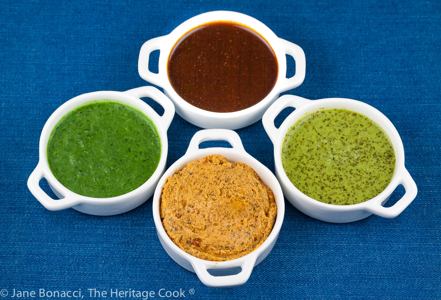 Mini white containers with four sauces for BBQ foods, two herbal sauces, a BBQ sauce, and a compound butter, all on a bright blue background; © 2023 Jane Bonacci, The Heritage Cook. 