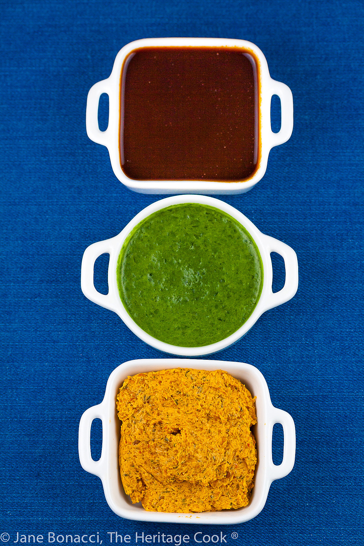 Mini white containers with four sauces for BBQ foods, two herbal sauces, a BBQ sauce, and a compound butter, all on a bright blue background; © 2023 Jane Bonacci, The Heritage Cook. 