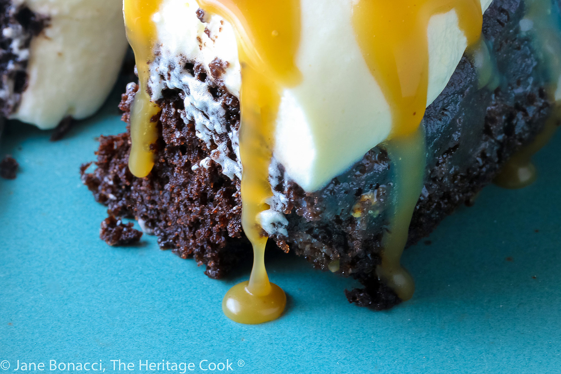 Focus on the caramel dripping off cake; A piece of chocolate cake studded with caramel bits and topped with pillowy whipped cream then sprinkled with mini chocolate chips and chopped pecans, on a turquoise plate © 2023 Jane Bonacci, The Heritage Cook. 