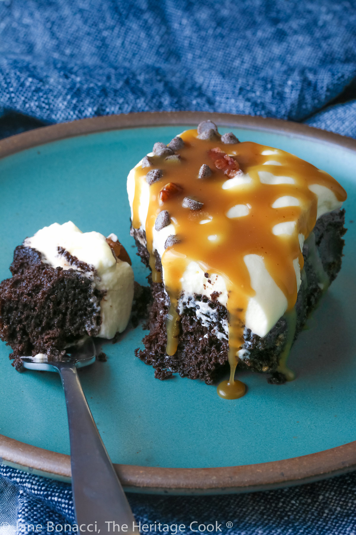 A piece of chocolate cake studded with caramel bits and topped with pillowy whipped cream then sprinkled with mini chocolate chips and chopped pecans, on a turquoise plate © 2023 Jane Bonacci, The Heritage Cook. 