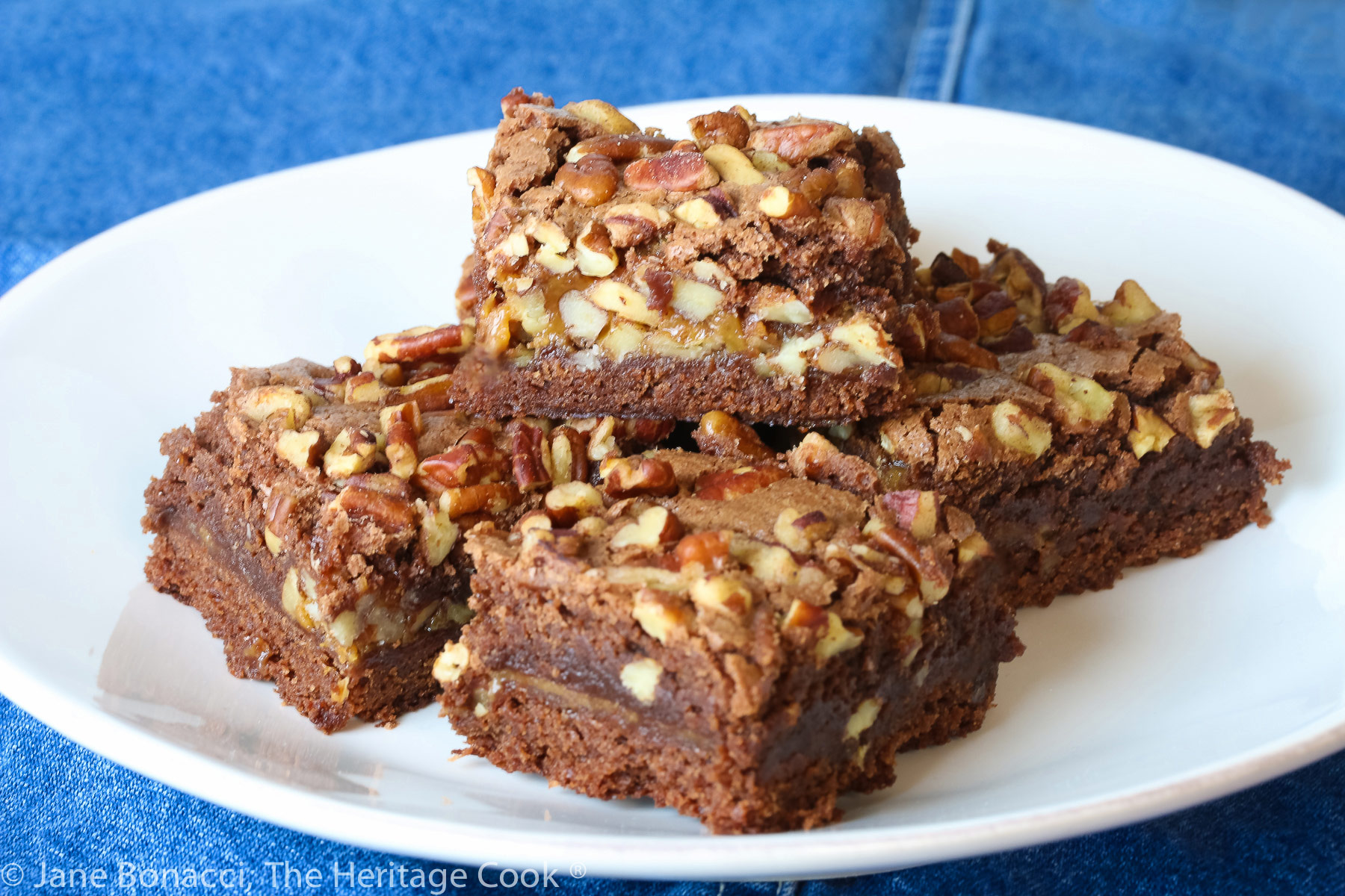 Stack of 5 brownies loaded with chopped pecans and caramel on white plate on blue denim napkin © 2023 Jane Bonacci, The Heritage Cook. 
