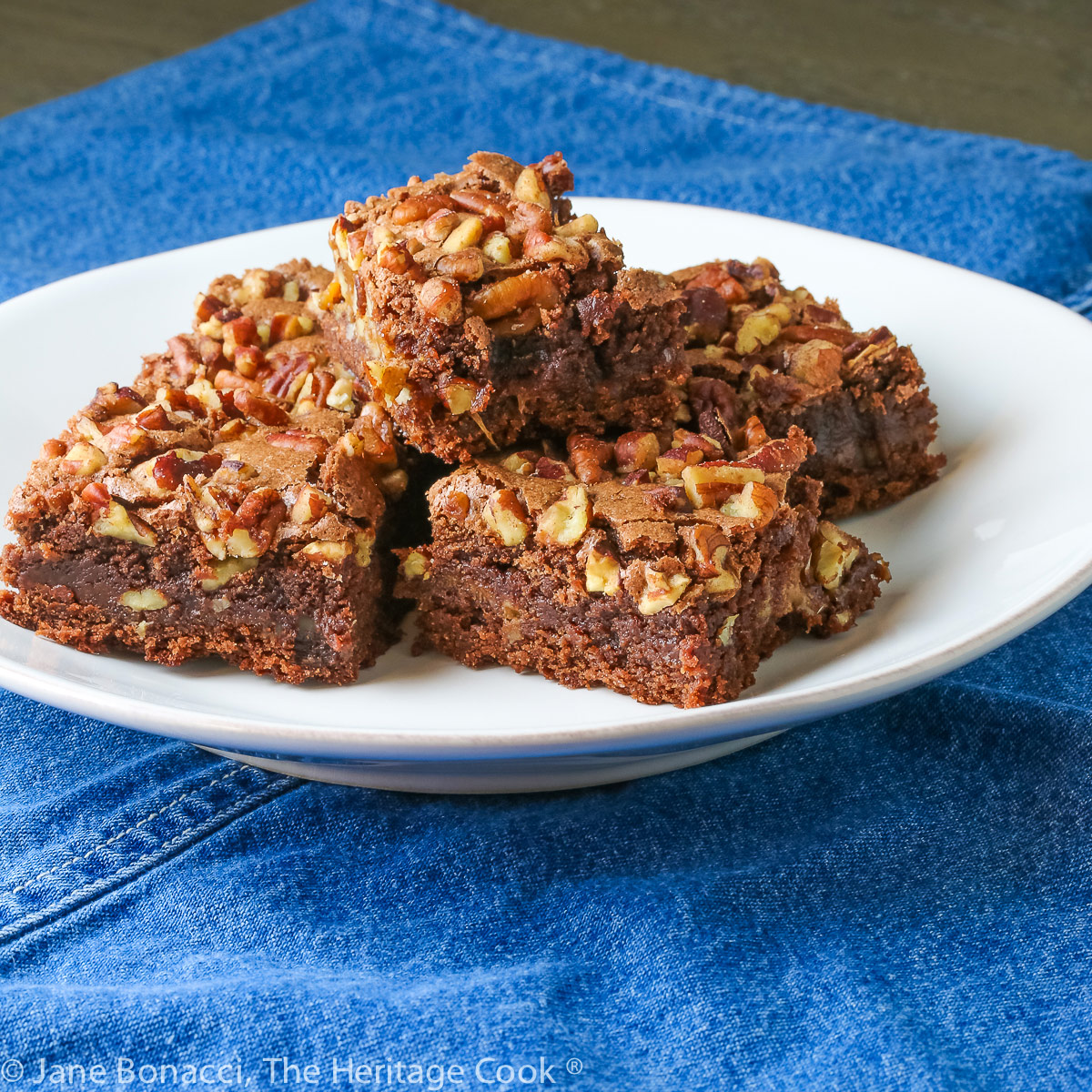 Stack of 5 brownies loaded with chopped pecans and caramel on white plate on blue denim napkin © 2023 Jane Bonacci, The Heritage Cook.