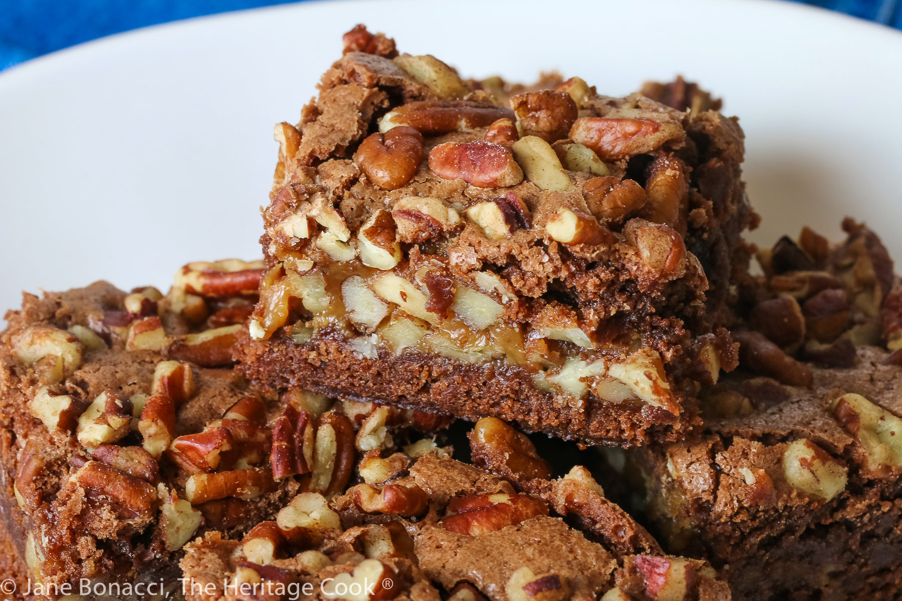 Stack of 5 brownies loaded with chopped pecans and caramel on white plate on blue denim napkin © 2023 Jane Bonacci, The Heritage Cook. 