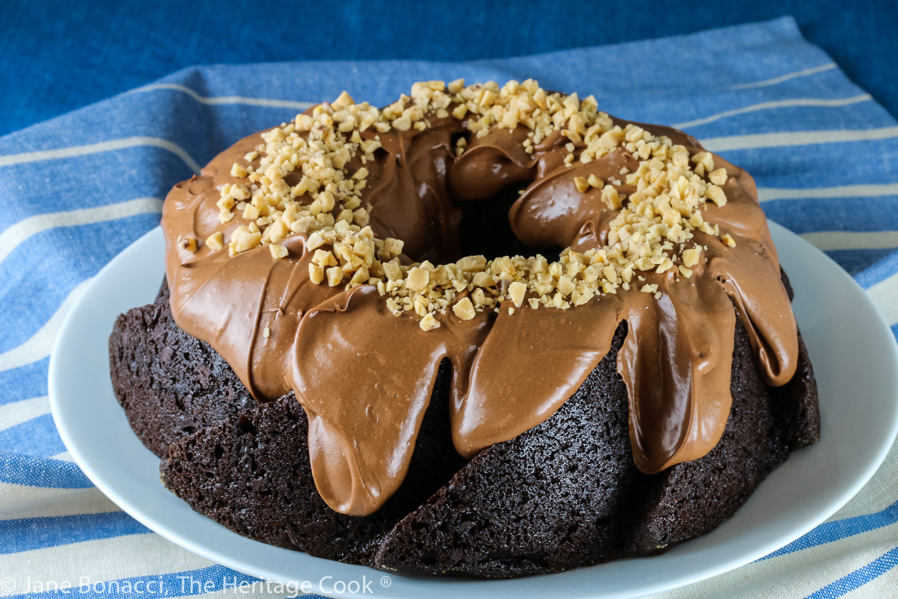 Swirl shaped bundt cake topped with fluffy chocolate frosting and toffee bits. Whole cake and slices on a white plate with blue striped cloth beneath © 2023 Jane Bonacci, The Heritage Cook. 