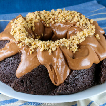Swirl shaped bundt cake topped with fluffy chocolate frosting and toffee bits. Whole cake and slices on a white plate with blue striped cloth beneath © 2023 Jane Bonacci, The Heritage Cook.