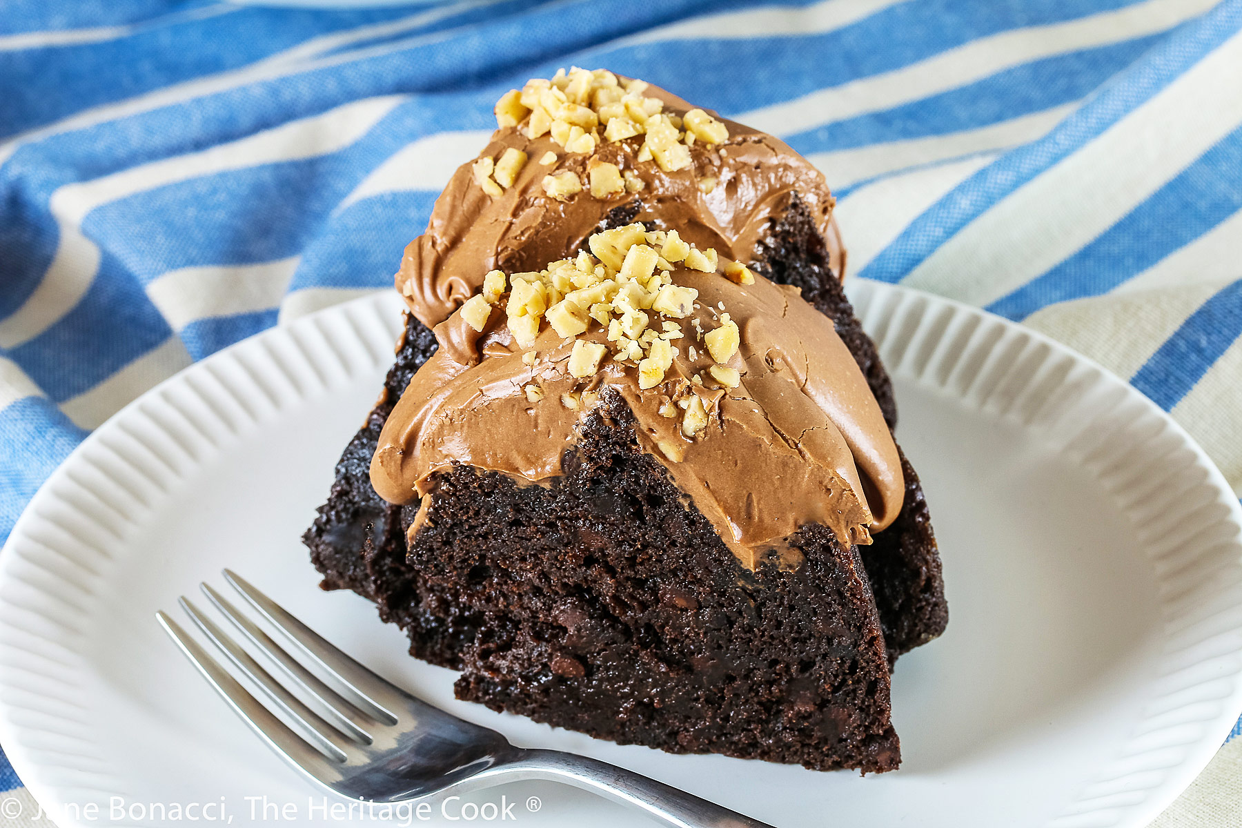 Swirl shaped bundt cake topped with fluffy chocolate frosting and toffee bits. Whole cake and slices on a white plate with blue striped cloth beneath © 2023 Jane Bonacci, The Heritage Cook. 