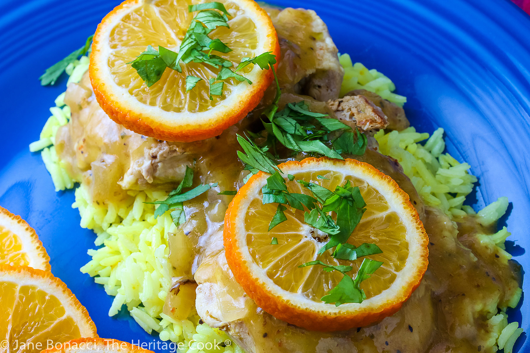 Two cooked chicken thighs resting on a pile of yellow rice on a blue plate, topped with the orange sauce, orange slices, chopped cilantro, and sliced almonds © 2023 Jane Bonacci, The Heritage Cook. 