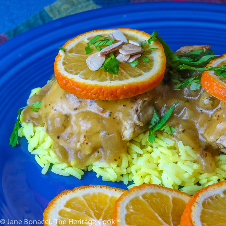 Two cooked chicken thighs resting on a pile of yellow rice on a blue plate, topped with the orange sauce, orange slices, chopped cilantro, and sliced almonds © 2023 Jane Bonacci, The Heritage Cook.