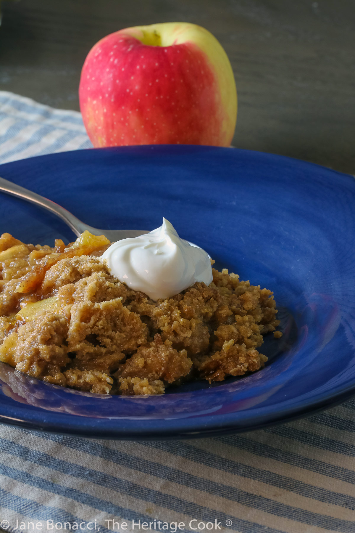 Scoop of apple crisp without oats in a wide, shallow dark blue bowl with a dollop of whipped cream on top; © 2023 Jane Bonacci, The Heritage Cook. 