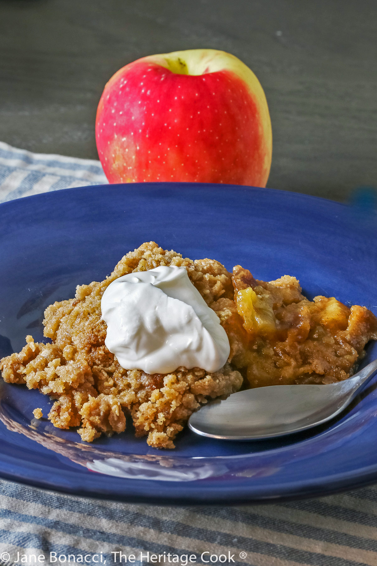 Scoop of apple crisp without oats in a wide, shallow dark blue bowl with a dollop of whipped cream on top; © 2023 Jane Bonacci, The Heritage Cook. 