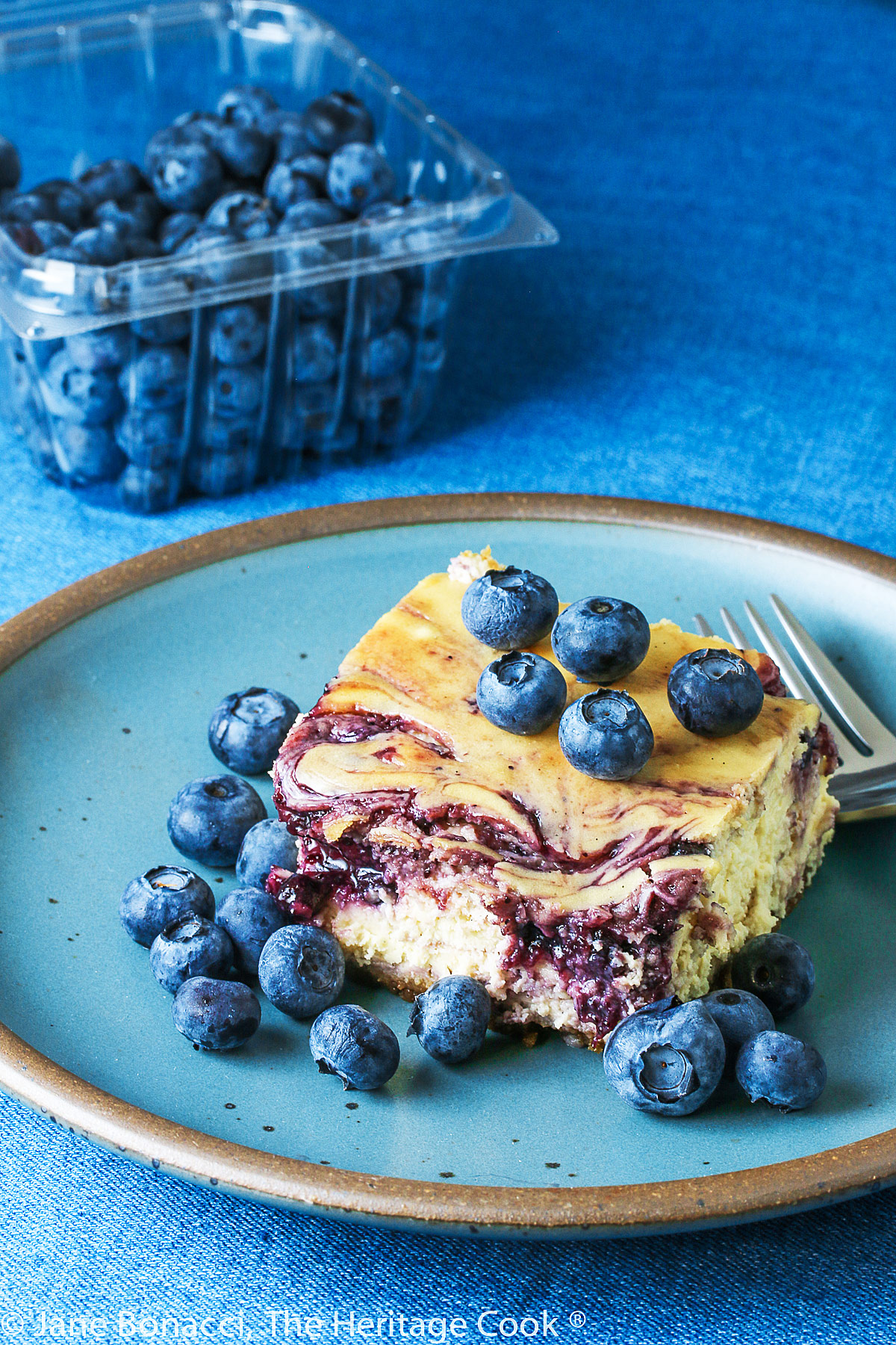 Square of Blueberry Swirl Cheese Cake Bars on turquoise plate on denim blue background surrounded by fresh blueberries © 2023 Jane Bonacci, The Heritage Cook. 