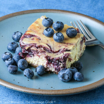 Square of Blueberry Swirl Cheese Cake Bars on turquoise plate on denim blue background surrounded by fresh blueberries © 2023 Jane Bonacci, The Heritage Cook.