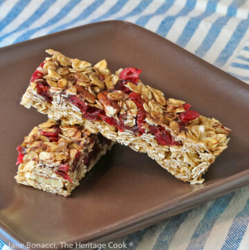 Two granola bars studded with red cranberries and chocolate set perpendicular to each other one on top in the shape of an X on a dark brown square plate © 2023 Jane Bonacci, The Heritage Cook.