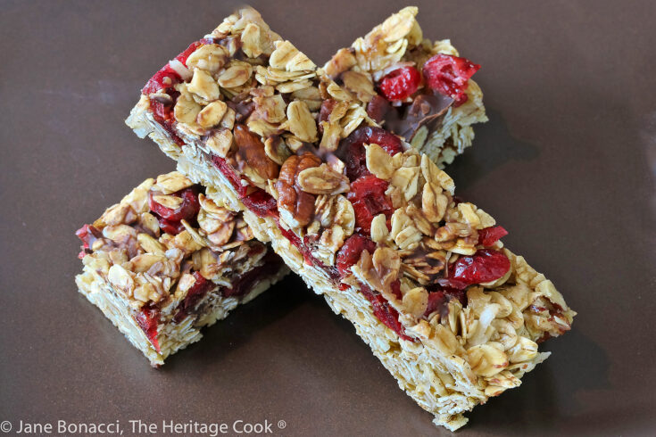 Two granola bars studded with red cranberries and chocolate set perpendicular to each other one on top in the shape of an X on a dark brown square plate © 2023 Jane Bonacci, The Heritage Cook.