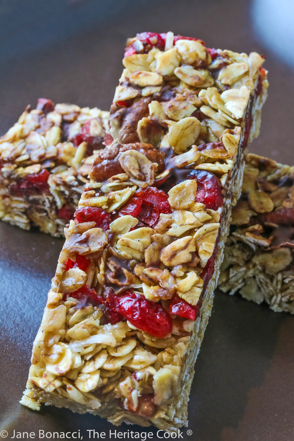 Two granola bars studded with red cranberries and chocolate set perpendicular to each other one on top in the shape of an X on a dark brown square plate © 2023 Jane Bonacci, The Heritage Cook. 